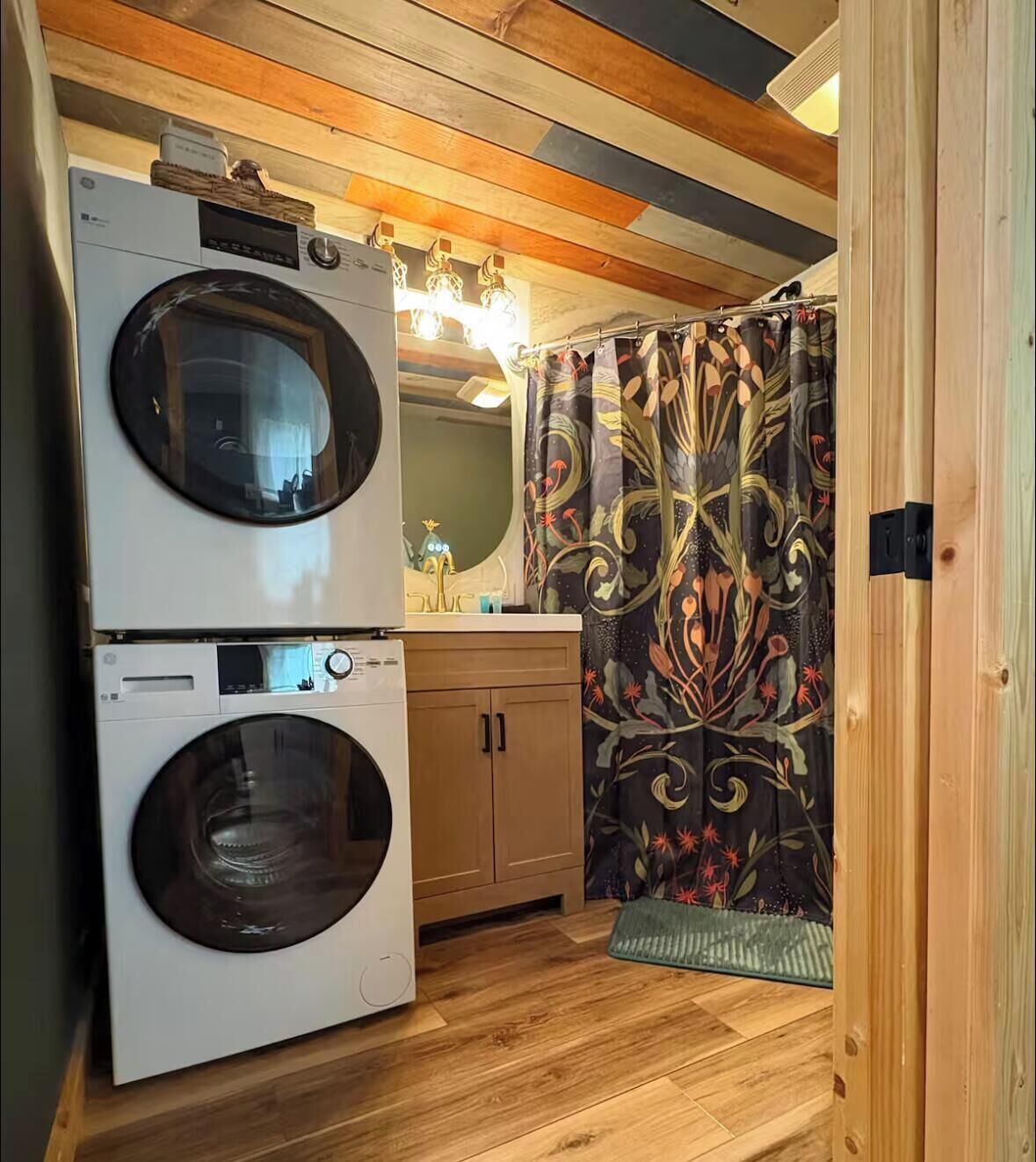 Une petite buanderie équipée de deux machines à laver superposées, de meubles en bois, d’un miroir et d’un rideau de douche à motifs.