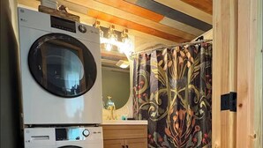 Een kleine wasruimte met twee gestapelde wasmachines, houten kasten, een spiegel en een douchegordijn met een patroon.