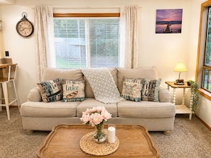 Un salon avec un canapé, des coussins décoratifs, une horloge, une fenêtre avec des stores, une lampe et un tableau encadré au mur.
