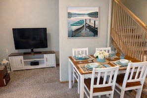 Un coin repas avec une table dressée pour quatre personnes, un téléviseur posé sur un meuble et un tableau représentant un lac.