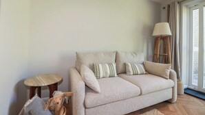 A neutral-toned sofa with striped pillows, a small round stool, a tall floor lamp, and a woven basket. - 67 Manor Villas (St. Columb Major)
