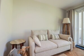 Ein sofa in neutralen Farbtönen mit gestreiften Kissen, ein kleiner runder Hocker, eine hohe Stehlampe und ein gewebter Korb.