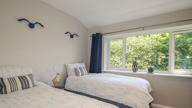A bedroom with two beds, a window with a view of trees, and a decorative bird on the wall. - 67 Manor Villas (St. Columb Major)