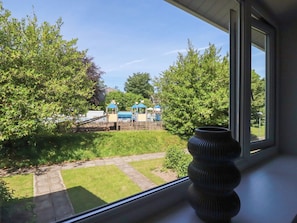 A view through a window of a backyard with a wooden fence, a grassy lawn, and a play area with a slide and climbing structures. - 67 Manor Villas (St. Columb Major)