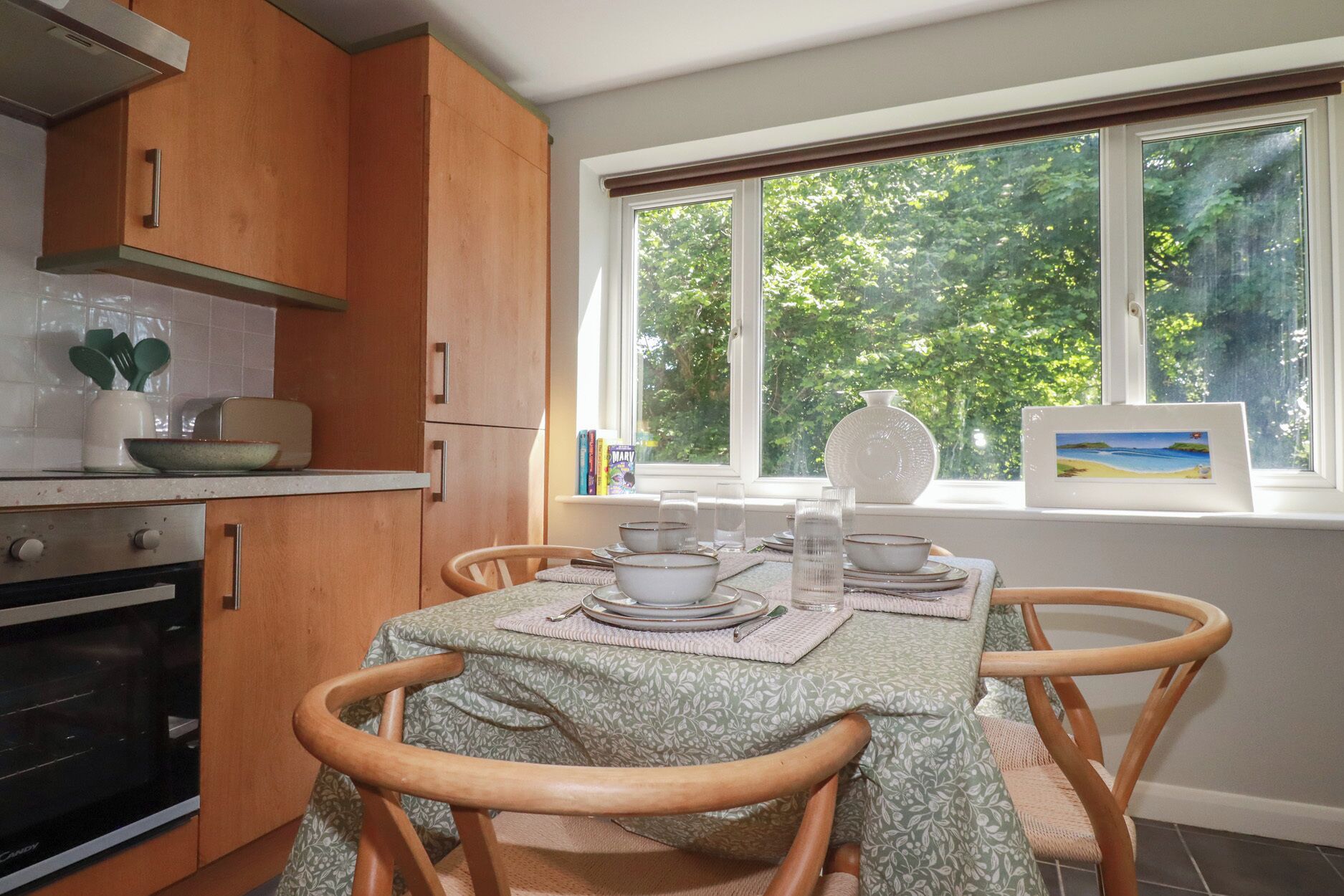 A kitchen with wooden cabinets, a dining table set with plates and glasses, and a window with a view of trees.
