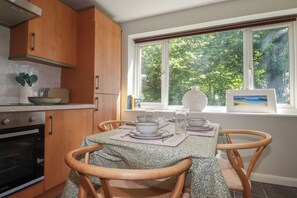 A kitchen with wooden cabinets, a dining table set with plates and glasses, and a window with a view of trees. - 67 Manor Villas (St. Columb Major)