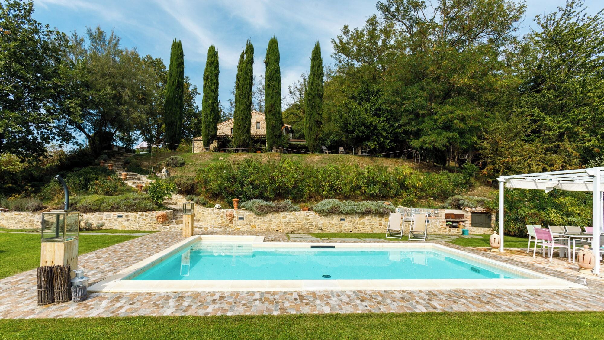 Una piscina con baldosas de piedra, una pérgola con sillas y un muro de piedra con esculturas.