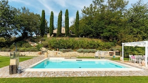 Ein Pool mit Steinfliesen, eine Pergola mit Stühlen und eine Steinsäule mit Skulpturen.