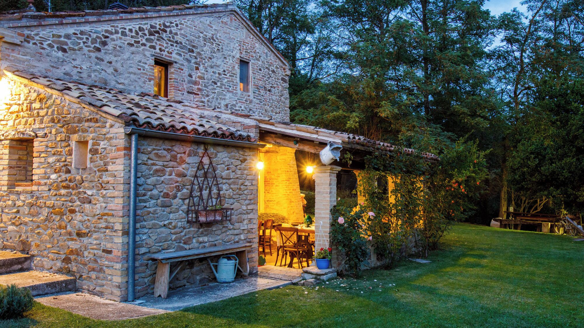 A stone building with a tiled roof, a wooden bench, and a table with chairs outside.