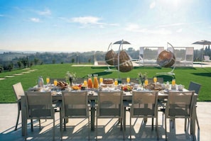 Un espace de restauration en plein air avec une table dressée pour le petit-déjeuner, entourée de chaises, et une vue panoramique en arrière-plan.