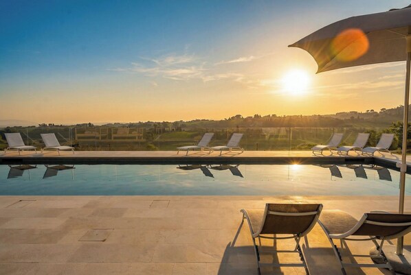 A pool with lounge chairs and an umbrella, overlooking a landscape with buildings and fields.