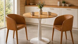 Una cocina moderna con una mesa redonda, dos sillas, un florero con flores, armarios de madera y una ventana con vista a árboles.