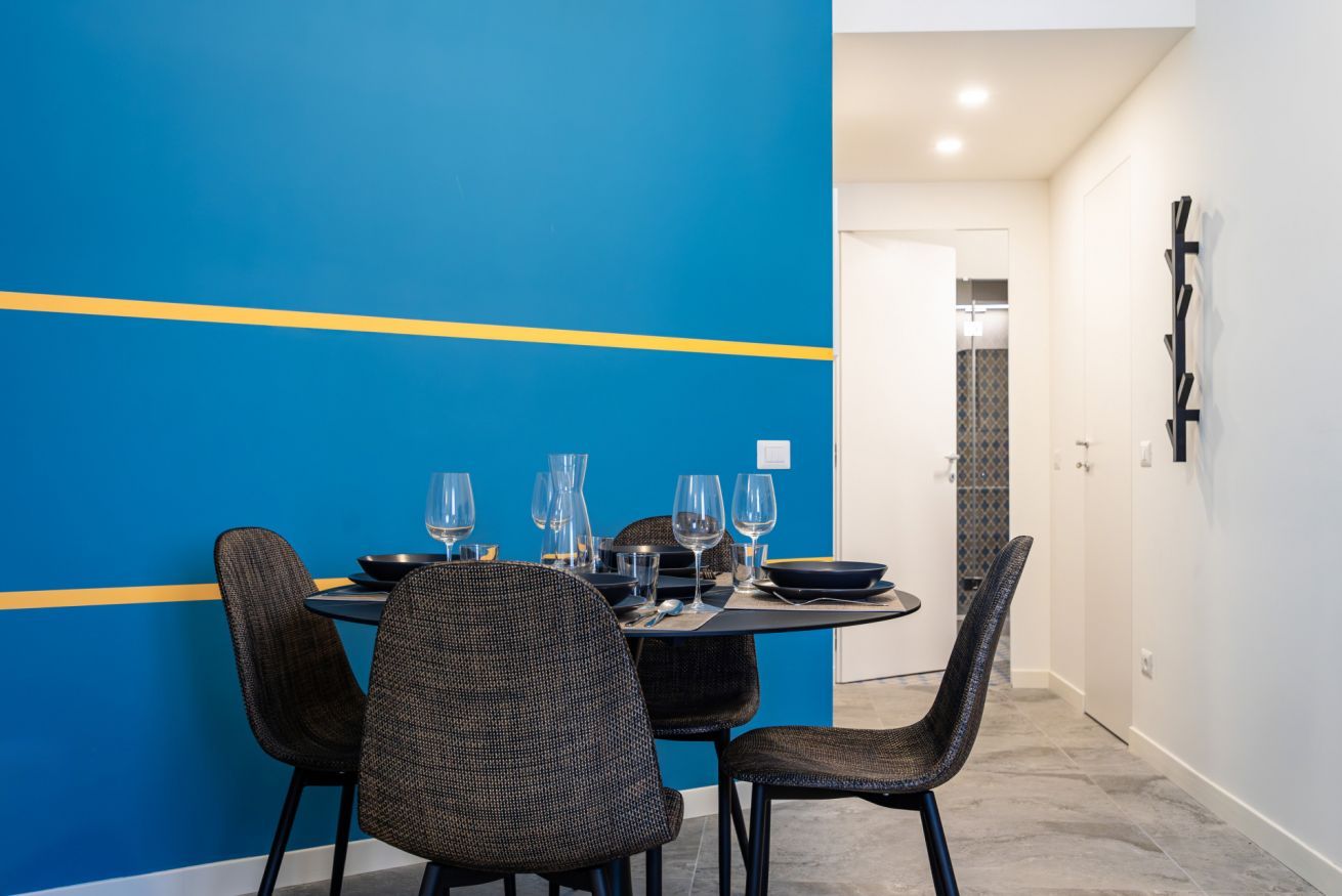 A dining area with a round table set for four, surrounded by chairs, in a room with a blue accent wall and a white door in the background.