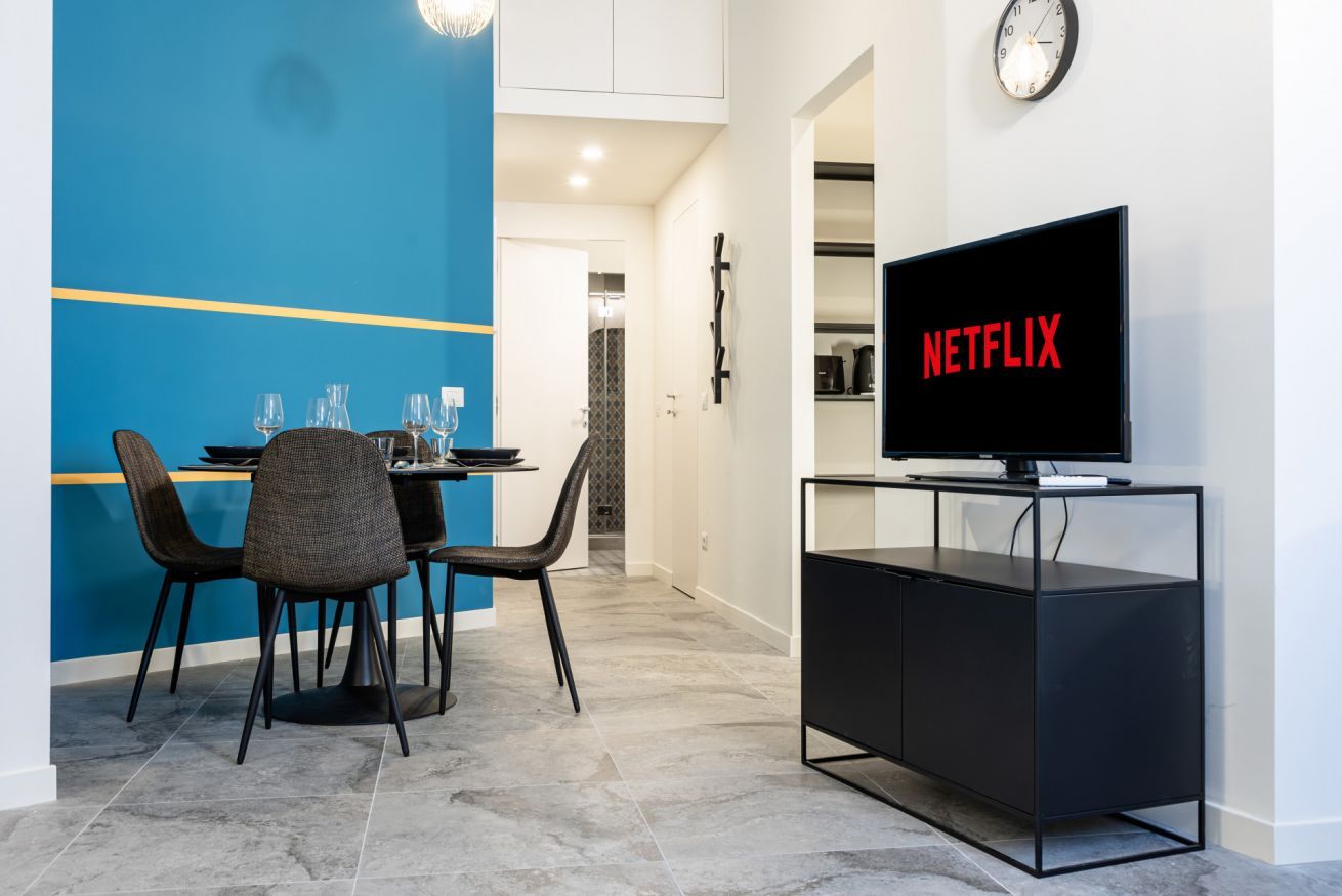 A modern living room with a dining area, a television on a stand, and a clock on the wall.