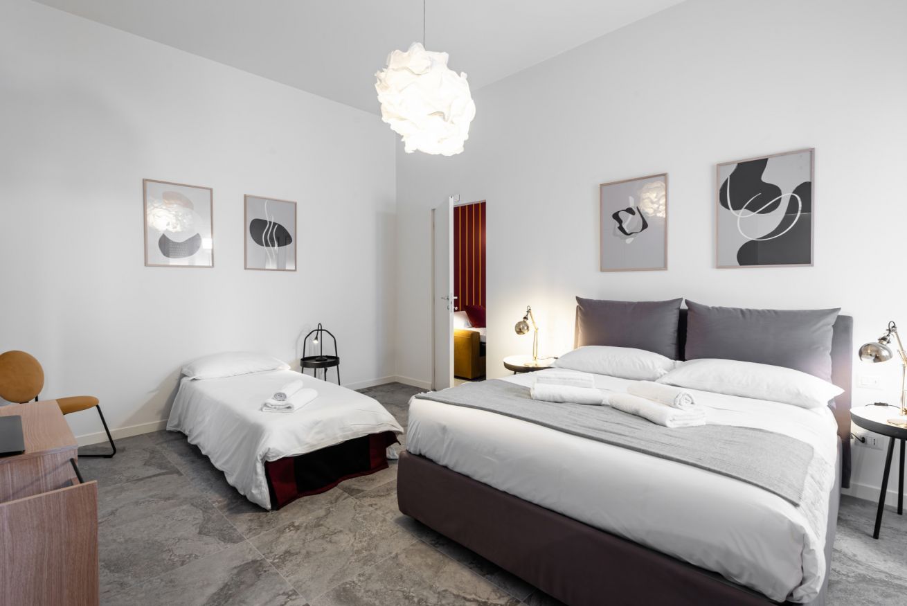 A modern hotel room with two beds, a desk, a chair, a hanging light fixture, and artwork on the walls.