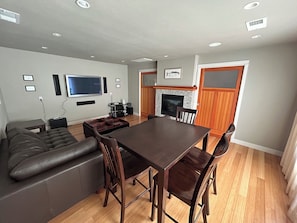 A living room with a dark brown dining table, wooden chairs, a leather sofa, a fireplace, a flat-screen TV mounted on the wall, and a wooden floor.