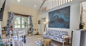 Ein Wohnzimmer mit Glastisch, weißen Stühlen, einer Couch mit Blattmuster, einem Gemälde an der Wand und einer Treppe.