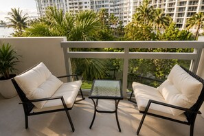 Een balkon met twee witte fauteuils met kussens en een glazen tafeltje, met uitzicht op een stadslandschap met palmbomen en een gebouw.