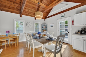 A dining room with a wooden table set for six, white chairs, a chandelier, and a kitchen area with white cabinets and a microwave. - East Dennis Gem! Walk to Beach! Sleeps 10! (Dennis)