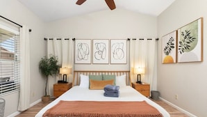 Un dormitorio con cama, dos mesitas de noche, un velador, ventilador de techo, ventana con persianas y obras de arte en las paredes.