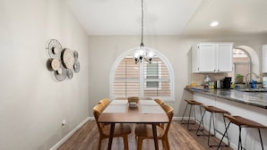 Un comedor con mesa y sillas de madera, una araña y una cocina con armarios blancos y un ventanal.