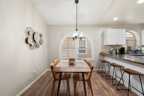 Un comedor con mesa y sillas de madera, una araña y una cocina con armarios blancos y un ventanal.