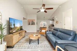 A modern living room with a flat-screen TV, a wooden entertainment unit, a blue leather sofa, a coffee table, and a ceiling fan. - Pool Home Near Cardinals | Great for Families (Peoria)