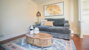 Ein Wohnzimmer mit grauem Sofa, einem runden Couchtisch, einem gemusterten Teppich und einem gerahmten Kunstwerk an der Wand.