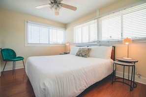A bedroom with a bed, a ceiling fan, two windows with blinds, a chair, and a bedside table with a lamp.