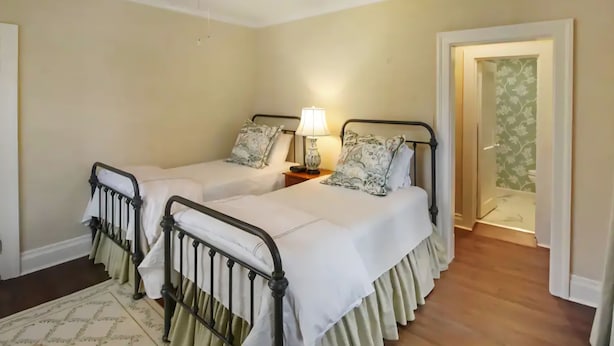 A bedroom with two beds, a nightstand with a lamp, a ceiling fan, and a door leading to another room.