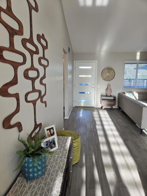 Ein modernes Wohnzimmer mit einem grauen Sofa, Wanddeko aus Holz, einer Topfpflanze und einem kleinen Tisch mit Bilderrahmen.