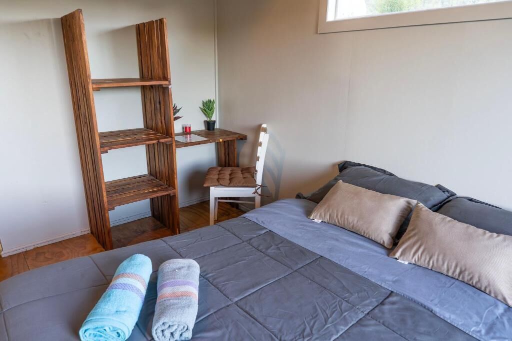 Un dormitorio con cama, almohadas, una silla, una mesita pequeña y una estantería de madera.