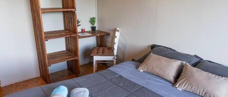 Un dormitorio con cama, almohadas, una silla, una mesita pequeña y una estantería de madera.