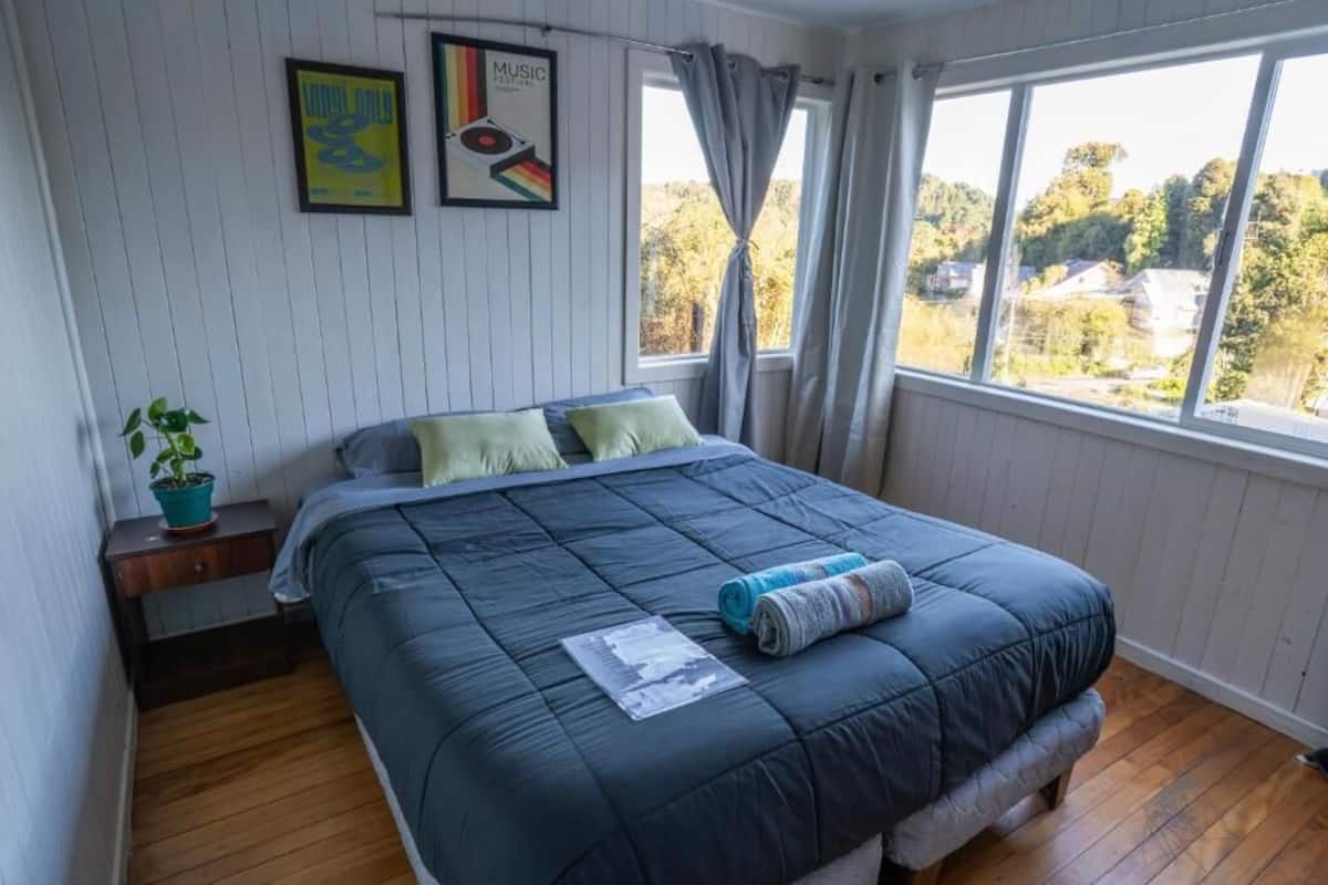 A bedroom with a bed, a nightstand, a potted plant, and a window with a view of houses and trees.