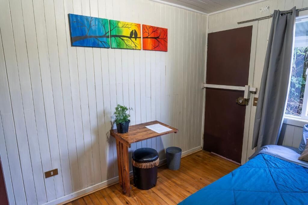 A room with a wooden desk, a small trash bin, a bed with a blue cover, and a door.