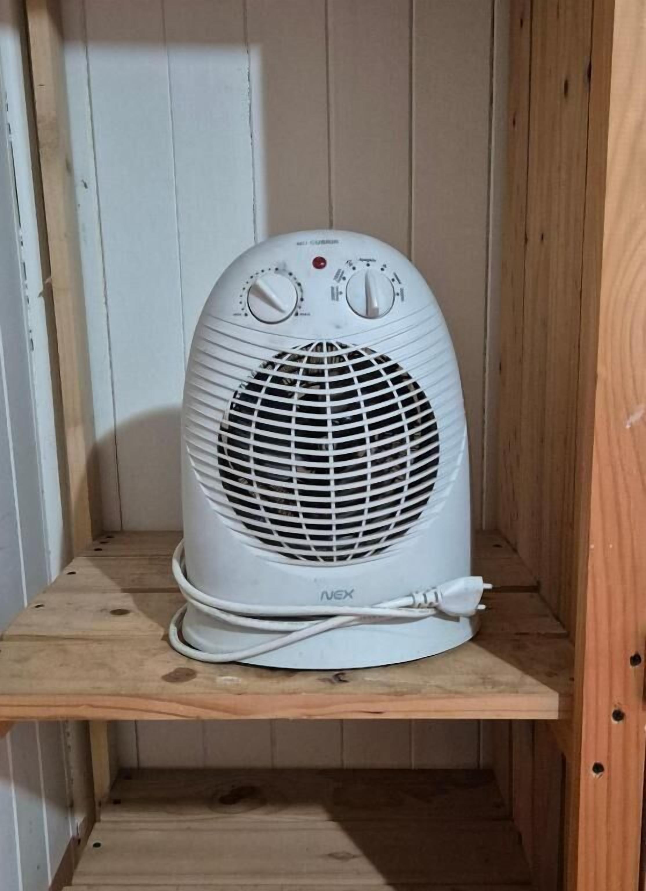 A white portable heater with control knobs and a power cord, placed on a wooden shelf in a room with wooden walls.