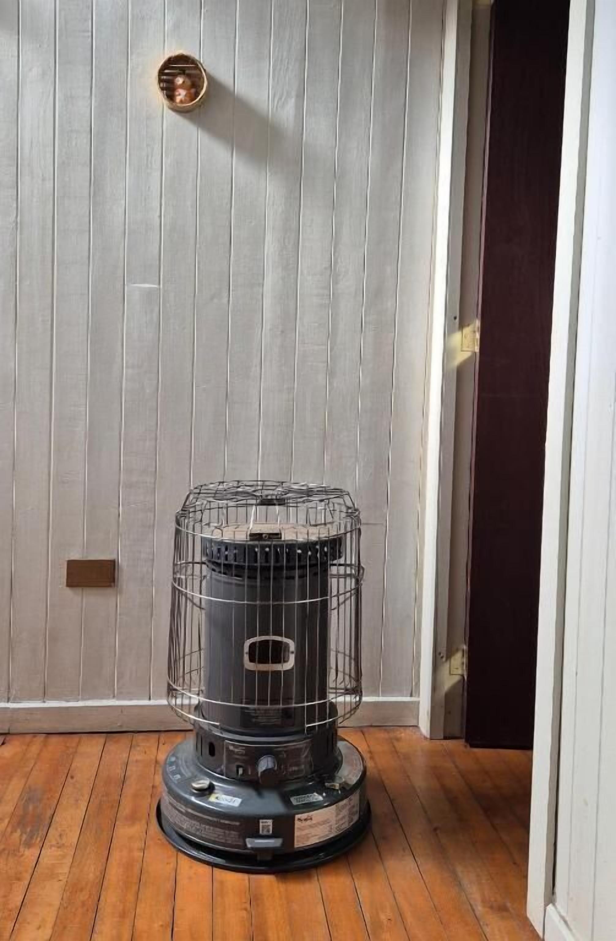 A portable heater in a room with wooden flooring and a white paneled wall.