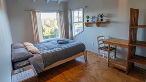 A bedroom with a bed, a wooden desk, a chair, a window with curtains, and a shelf with plants. - hostal refugio nomada (Puerto Montt)