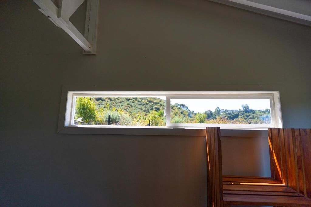 Een kamer met een raam dat uitkijkt op een groen landschap, een houten bankje en een witte balk.
