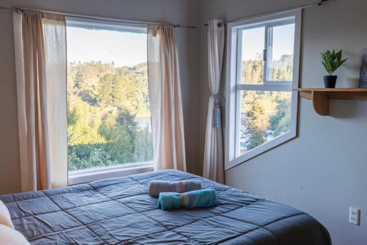 A bedroom with a bed, two windows with curtains, a view of trees, and a small shelf with a plant.