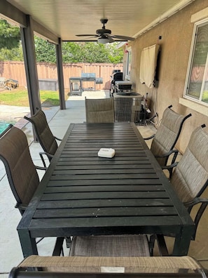 Eine überdachte Terrasse mit Tisch, Stühlen, einem Deckenventilator und einem Grill im Hintergrund.
