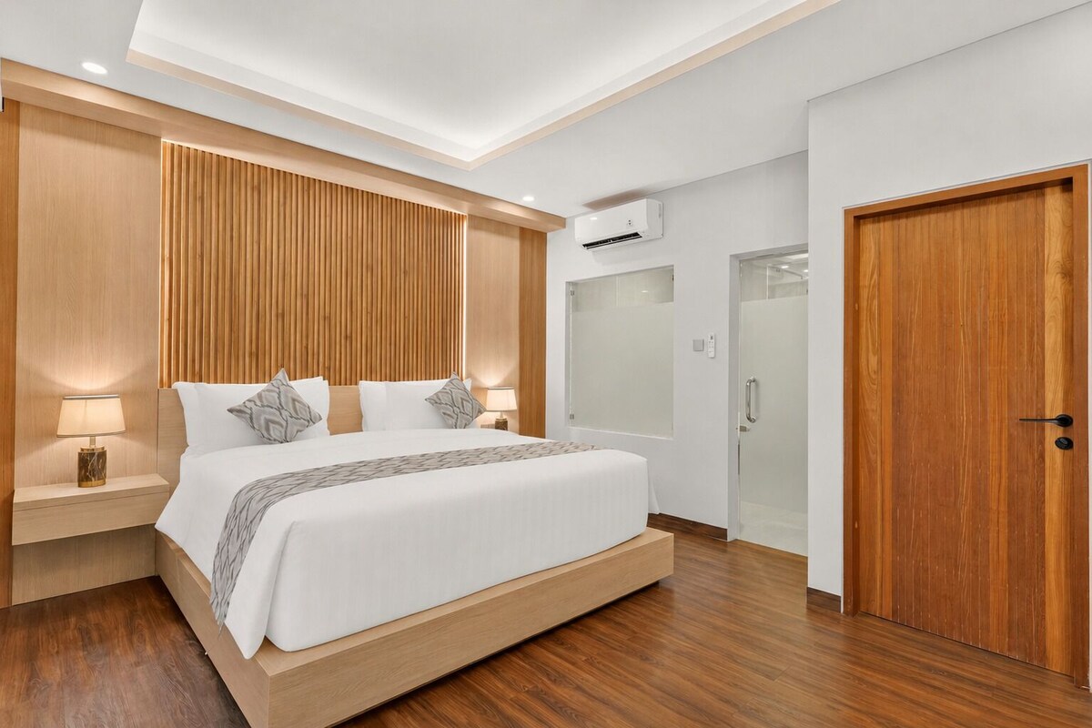 A modern hotel room with a wooden headboard, a large bed, bedside lamps, an air conditioning unit, a glass shower enclosure, and a wooden door.