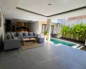 En moderne stue med en grå sofa, et sofabord og et poolområde med planter.