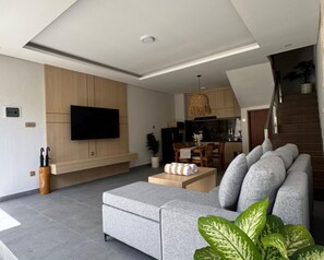 A modern living room with a grey sofa, a flat-screen TV mounted on the wall, a dining area with wooden cabinets, and a staircase leading to an upper level. - The Radiance Villas Seminyak (Badung)