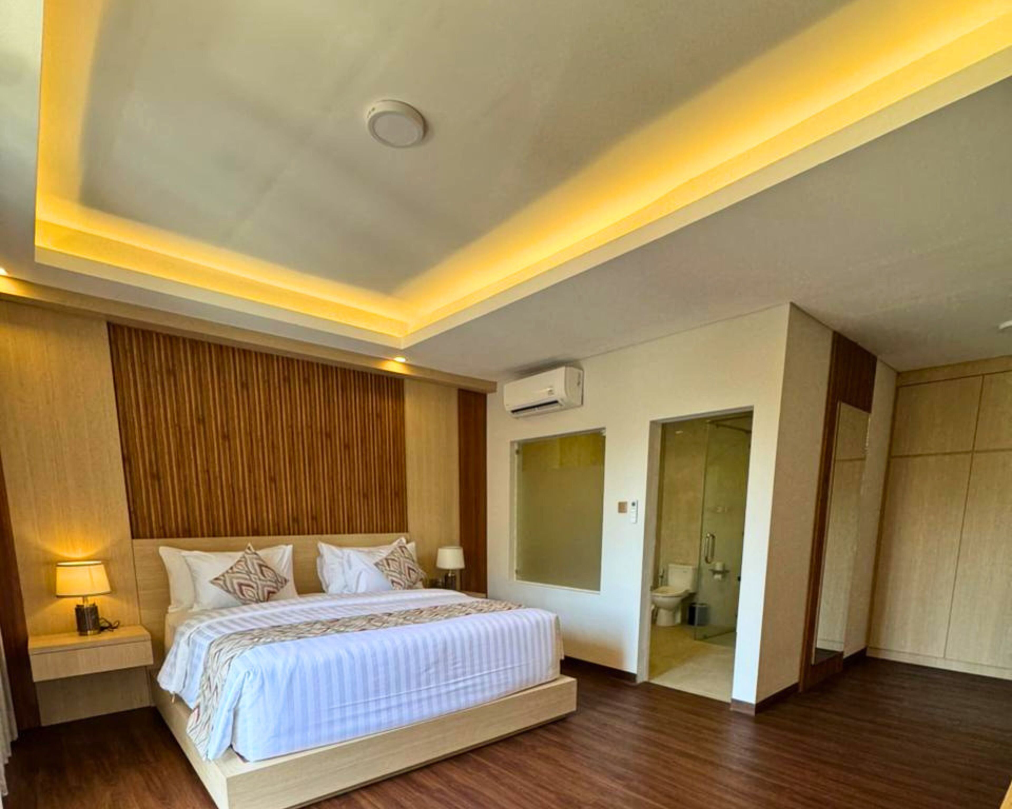 Ein modernes Hotelzimmer mit einem großen Bett, Nachttischen, einem Kleiderschrank und einem durch eine geöffnete Tür einsehbaren Badezimmer.