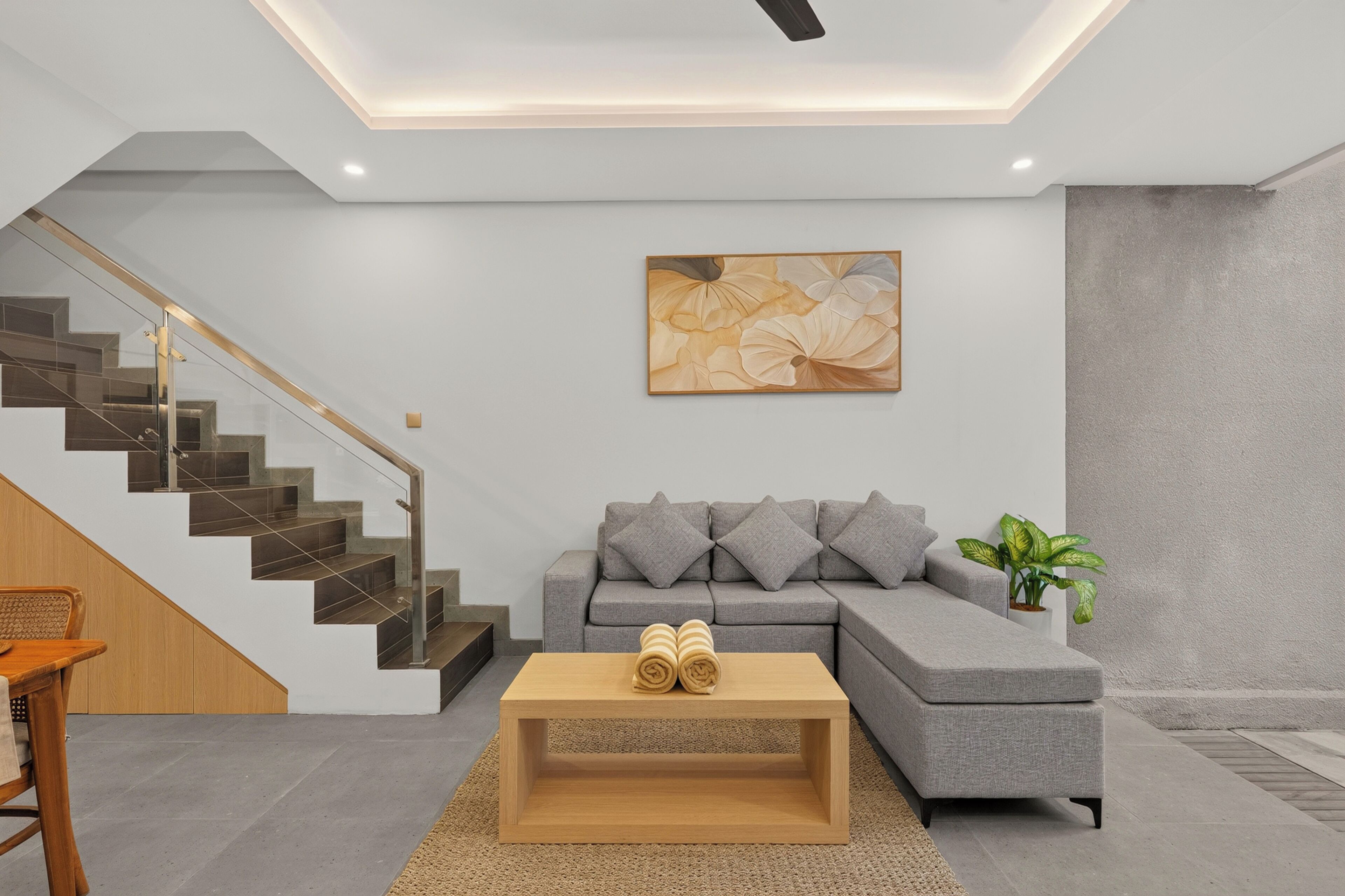 A modern living room with a grey sectional sofa, a wooden coffee table, a staircase with glass railings, and a painting on the wall.