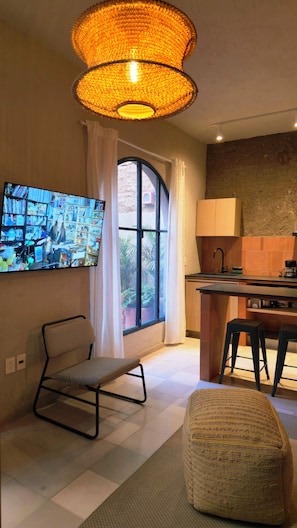 A modern living room with a television, a comfortable chair, a sofa, a kitchen area, and a large window with curtains. - Loft Chic 2 Niveles Red Descanso Rooftop Grill Catedral Santuario S3 (Guadalajara)