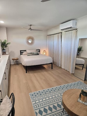 A bedroom with a bed, a ceiling fan, an air conditioning unit, a bedside lamp, a small table with a chair, and a mirror. - Private beautiful modern 400 sq ft studio located in a quiet neighborhood in SCW (Sun City West)