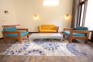 Une pièce avec un canapé jaune, deux chaises en bois, une petite table, des tapis à motifs et des lampes fixées au mur.