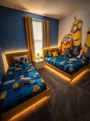 Ein Schlafzimmer mit zwei Betten, jeweils mit blauen Laken mit Minion-Motiven, einem großen Minion-Wandgemälde und einem Fenster mit gelben Vorhängen.
